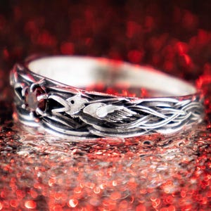 Gothic sterling silver raven ring with braided feather band and deep red garnet gemstone, photographed against a dramatic red bokeh background