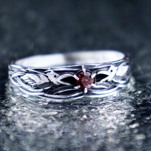 Handcrafted 925 sterling silver Huginn and Muninn raven ring with round-cut garnet, displayed on a wet dark stone surface with soft reflections