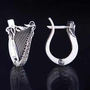 May include: A pair of silver-colored hoop earrings. One earring features a detailed harp design with intricate lines and a woven pattern. The other earring has a simple, curved design with a clasp.