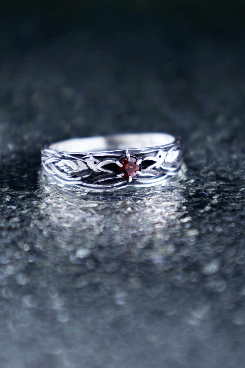 Handcrafted 925 sterling silver Huginn and Muninn raven ring with round-cut garnet, displayed on a wet dark stone surface with soft reflections