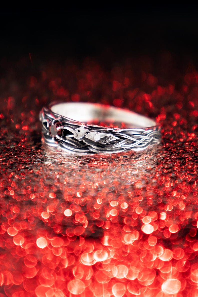 Gothic sterling silver raven ring with braided feather band and deep red garnet gemstone, photographed against a dramatic red bokeh background