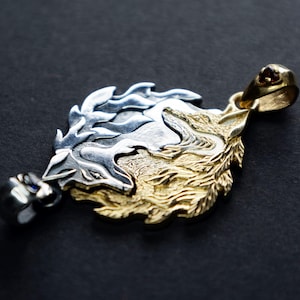 Puede incluir: Un colgante de plata y oro con un diseño detallado de dos lobos enfrentados. El colgante tiene un bucle en la parte superior para sujetarlo a una cadena.