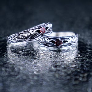Two sterling silver Norse raven rings with garnet stones displayed together on a reflective wet stone surface, showing both narrow and wide band variants as a matching couple set