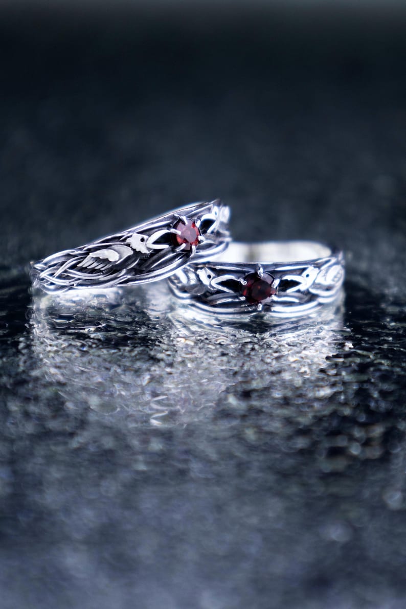 Two sterling silver Norse raven rings with garnet stones displayed together on a reflective wet stone surface, showing both narrow and wide band variants as a matching couple set