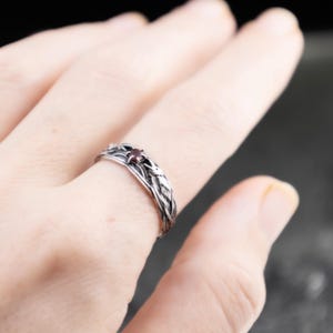Gothic sterling silver raven ring with red garnet worn on a woman's middle finger, feather braid detail visible against a dark background