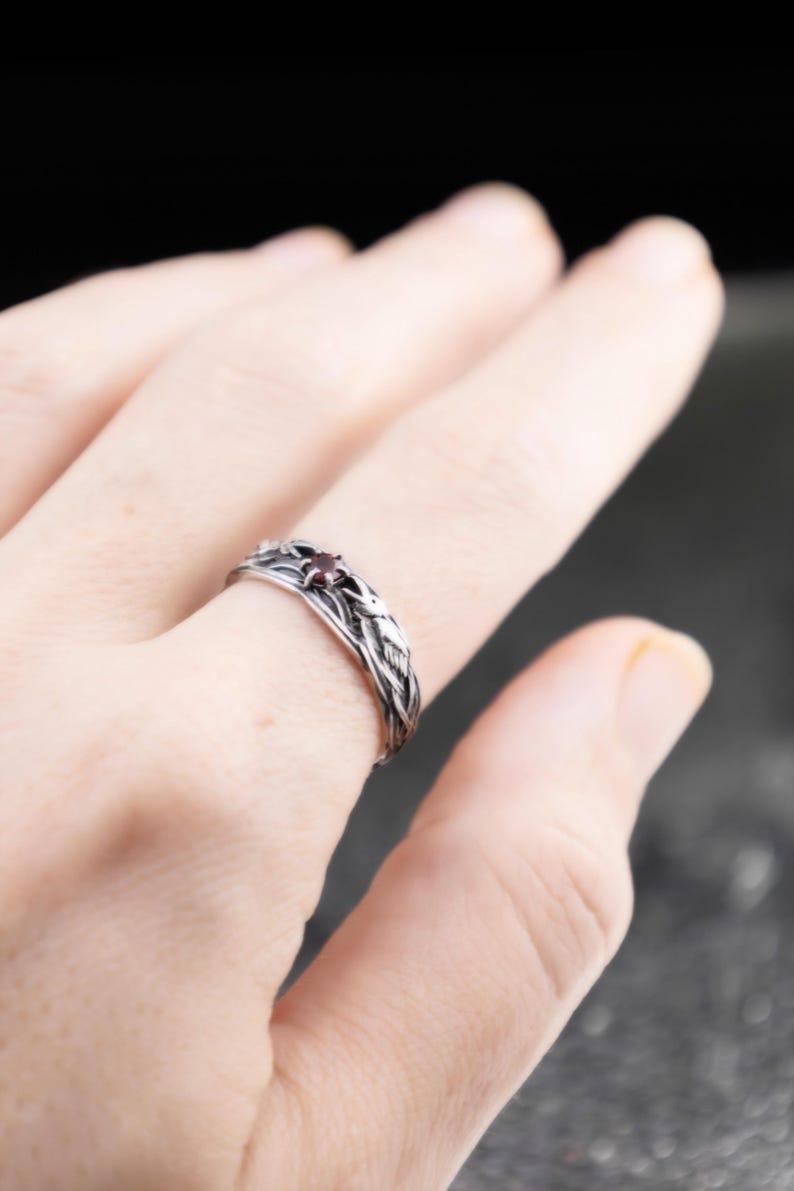 Gothic sterling silver raven ring with red garnet worn on a woman's middle finger, feather braid detail visible against a dark background