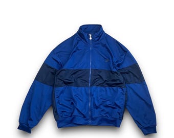 May include: A blue track jacket with a full zipper and a high collar. The jacket features a dark blue horizontal stripe across the chest and upper back. A small logo is visible on the upper left chest.
