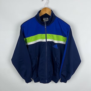 Puede incluir: Una chaqueta de chándal Adidas azul marino con una franja verde y blanca en el pecho. El logotipo de Adidas está bordado en el pecho izquierdo.