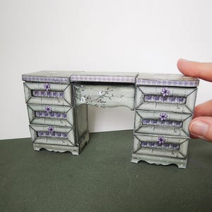 May include: A miniature, light green desk with three drawers on each side, adorned with purple floral accents. The desk features a decorative floral design on the front and a scalloped edge. The desk is a charming piece of dollhouse furniture.