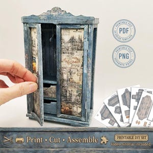 Puede incluir: Un pequeño armario azul en miniatura con puertas abiertas, que revela una percha y cajones. El armario está decorado con estampados arquitectónicos vintage. La imagen incluye el texto "Print Cut Assemble" y "Printable DIY Set".