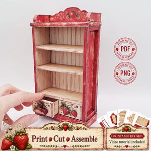 Op de afbeelding: Een miniatuur houten rek met een verweerde rode afwerking, voorzien van drie open planken en twee kleine lades onderaan. Het meubel is versierd met aardbei-illustraties en bevat de tekst Print Cut Assemble.