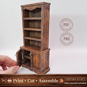 Op de afbeelding: Een kleine houten kast met een verweerde afwerking. De kast heeft een buffetkast met planken en een lager gedeelte met een deur op een kier. De tekst "Print Cut Assemble" staat onderaan, samen met "Printable DIY Set".
