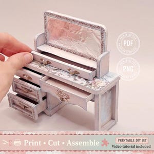 Puede incluir: Una miniatura de tocador blanco con espejo, cajones y detalles florales. El tocador tiene un aspecto envejecido y vintage. El texto "Print Cut Assemble" está en la parte inferior. Es un kit de bricolaje imprimible.