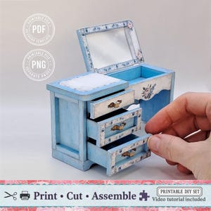 May include: A miniature blue dresser with three drawers, a mirror, and floral accents. The text "Print Cut Assemble" is displayed below the dresser. The image also includes the text "Printable DIY Set" and "Video tutorial included".