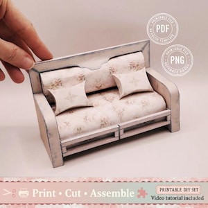 Op de afbeelding: Een miniatuur DIY-bank met een bloempatroon zitting en twee bijpassende kussens. De bank is lichtroze en wit, met de tekst "Print Cut Assemble" onderaan. Inclusief afdrukbare PDF- en PNG-bestanden.