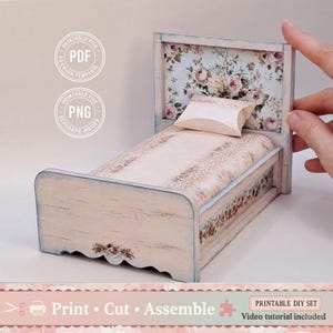Puede incluir: Una cama de muñecas en miniatura con cabecero floral y ropa de cama a juego. El marco de la cama está pintado de blanco con detalles en azul desgastado. El texto "Print Cut Assemble" es visible.
