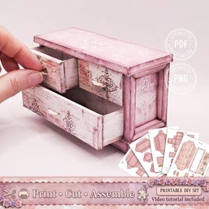 Op de afbeelding: Een miniatuur roze en witte decoratieve ladekast met twee open lades. De kast heeft een verweerde afwerking en sierlijke details. De afbeelding bevat de tekst "Print Cut Assemble" en "Printable DIY Set".