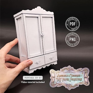 May include: A white paper DIY wardrobe with two doors and two drawers, held by a hand. The wardrobe has decorative handles and a scalloped base. Text on the image includes "PRINTABLE DIY SET" and "Video tutorial included."
