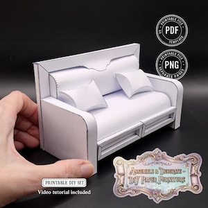 May include: A white papercraft sofa with two matching pillows. The DIY furniture set includes a PDF template and a video tutorial. The sofa is held by a hand, showcasing the scale of the miniature furniture.