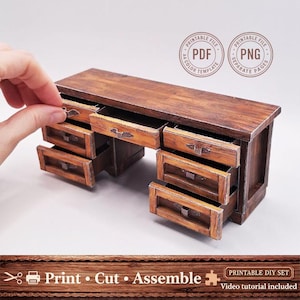 May include: A miniature wooden desk with multiple drawers, some open, showcasing a detailed, aged finish. The desk has metal handles and is part of a printable DIY set. The text "Print Cut Assemble" is visible.
