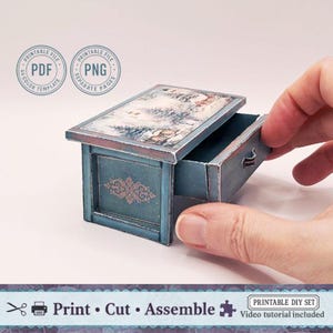 May include: A small blue chest of drawers with a landscape scene on the lid. The chest has an ornate design on the front and is being opened by a hand. The image includes text that says "Print, Cut, Assemble".