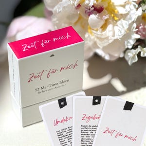 Könnte beinhalten: Eine weiße rechteckige Schachtel mit einem leuchtend pinken Deckel und der Aufschrift Zeit für mich. Die Box ist mit 52 Me-Time Ideen für bewusste Auszeiten beschriftet. Drei weiße Karten mit schwarzen Symbolen und rotem Text liegen davor.