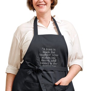 May include: A black apron with a white quote that reads "A feast is made for laughter, wine makes life merry, and money is the answer for Ecclesiastes 10:19".