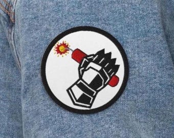 May include: A circular embroidered patch featuring a black armored gauntlet holding a red dynamite stick with a lit yellow and orange fuse. The patch has a white background and a thick black border, attached to blue denim fabric.
