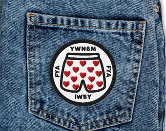 May include: A circular embroidered patch featuring a white background with a black border. The center displays a pair of white shorts patterned with small red hearts. The text YWNBM, IWBY, and FYA is stitched in black around the shorts.