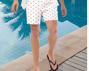 May include: White swim trunks featuring a repeating pattern of small red hearts. The shorts have an elastic waistband and a relaxed fit, extending to mid-thigh. The fabric is smooth and lightweight, designed for water activities and casual wear.
