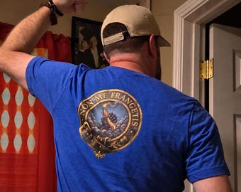 May include: A blue t-shirt featuring a circular graphic on the back. The design depicts a gauntleted hand holding a flaming torch amidst clouds, surrounded by a belt-like border with the text "NON ME FRANGETIS" in gold lettering.