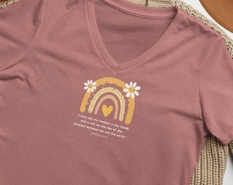 May include: Dusty rose v-neck t-shirt featuring a rainbow design with daisies and a heart. The rainbow is yellow, orange, and brown. The text reads, "I have set my rainbow in the clouds and it will be the sign of the covenant between me and the earth."