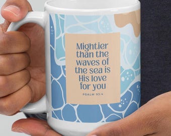 May include: White ceramic mug with a blue and tan abstract design. The mug features the text "Mightier than the waves of the sea is His love for you" from Psalm 93:4. The mug is held by a person.