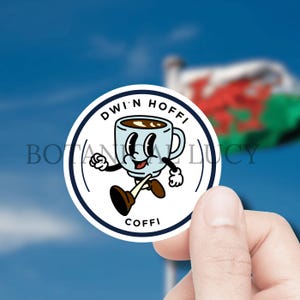 May include: A white round sticker with a blue border. The sticker features a cartoon coffee cup with a smiling face, walking and holding a cup of coffee. The text "Dwi'n Hoff" is above the cup and "Coffe" is below the cup.
