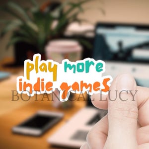 May include: A white sticker with the text "Play more indie games" in orange, yellow, and teal lettering. The sticker is being held by a hand.