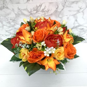 May include: A colourful floral arrangement with orange roses, lilies, and other flowers. The bouquet includes green leaves and small white flowers, creating a full, vibrant display. The flowers are arranged in a low, wide shape.