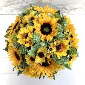 May include: A vibrant, round floral arrangement featuring bright yellow sunflowers with dark brown centers. The bouquet is accented with green succulents and foliage, creating a full, textured display. The arrangement is set against a white background.