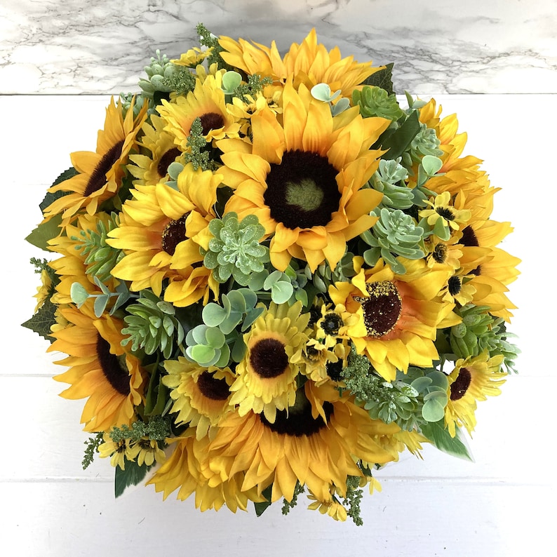 May include: A vibrant, round floral arrangement featuring bright yellow sunflowers with dark brown centers. The bouquet is accented with green succulents and foliage, creating a full, textured display. The arrangement is set against a white background.