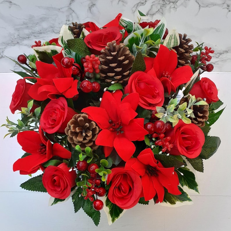 May include: A festive arrangement of red poinsettias, roses, and berries, accented with pine cones and green foliage. This holiday-themed floral display is perfect for seasonal decor, adding a touch of warmth and cheer to any space.