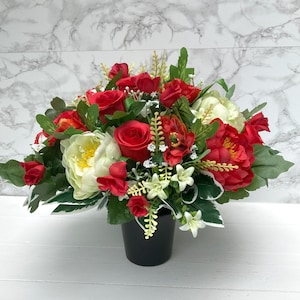 May include: A red and white artificial flower arrangement in a black pot. The arrangement features roses, peonies, and other flowers.