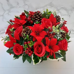 May include: A festive floral arrangement featuring vibrant red poinsettias, roses, and berries, accented with pinecones and lush green foliage. This decorative centerpiece is perfect for holiday decor.