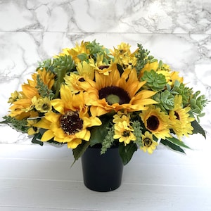 May include: A vibrant arrangement of artificial sunflowers, accented with green succulents and foliage, presented in a black pot. The sunflowers have bright yellow petals and dark brown centers, creating a cheerful and natural aesthetic.