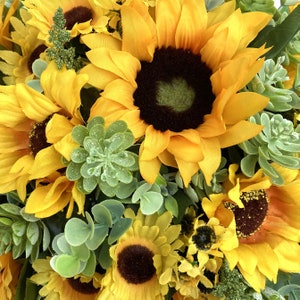 May include: Close-up of a vibrant floral arrangement featuring bright yellow sunflowers with dark brown centers. The arrangement includes green succulents and foliage, creating a natural and colorful display. The flowers are in full bloom.