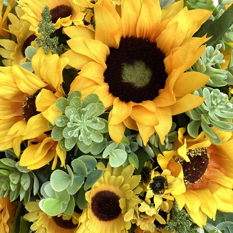 May include: Close-up of a vibrant floral arrangement featuring bright yellow sunflowers with dark brown centers. The arrangement includes green succulents and foliage, creating a natural and colorful display. The flowers are in full bloom.