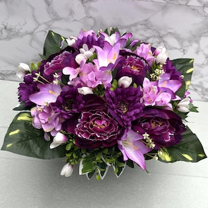 May include: A colourful floral display with a variety of purple, lilac, and white flowers. The bouquet includes large, deep purple peonies, elegant lilies, and smaller blooms, complemented by green foliage. The arrangement is set against a marble backdrop.
