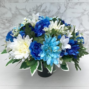 May include: A blue and white artificial flower arrangement in a black pot. The arrangement features blue roses, white daisies, and light blue flowers.