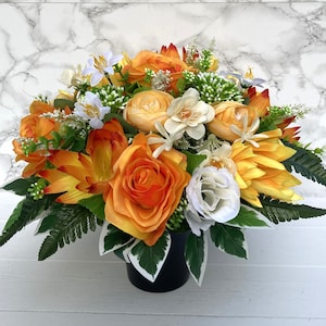 May include: A bouquet of artificial flowers in a black pot. The flowers are a mix of orange, yellow, and white. The bouquet is arranged in a circular shape.