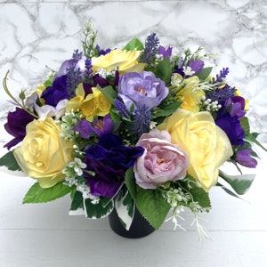 May include: A bouquet of yellow, purple and pink flowers arranged in a black vase. The flowers are a mix of roses, lilies and other blooms.
