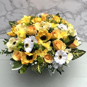 May include: A colourful floral display with yellow roses, anemones, and other flowers. The bouquet includes white and yellow blooms with green foliage, creating a bright and cheerful arrangement. The display is set against a neutral background.