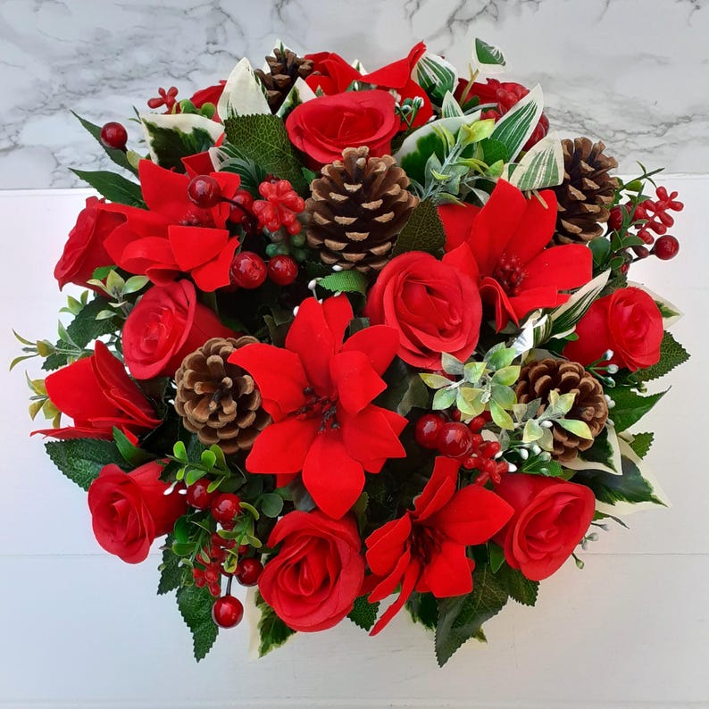 May include: A circular floral arrangement featuring vibrant red roses, poinsettias, and berries, interspersed with green foliage and pinecones. The arrangement is set against a white background, creating a festive and decorative display.